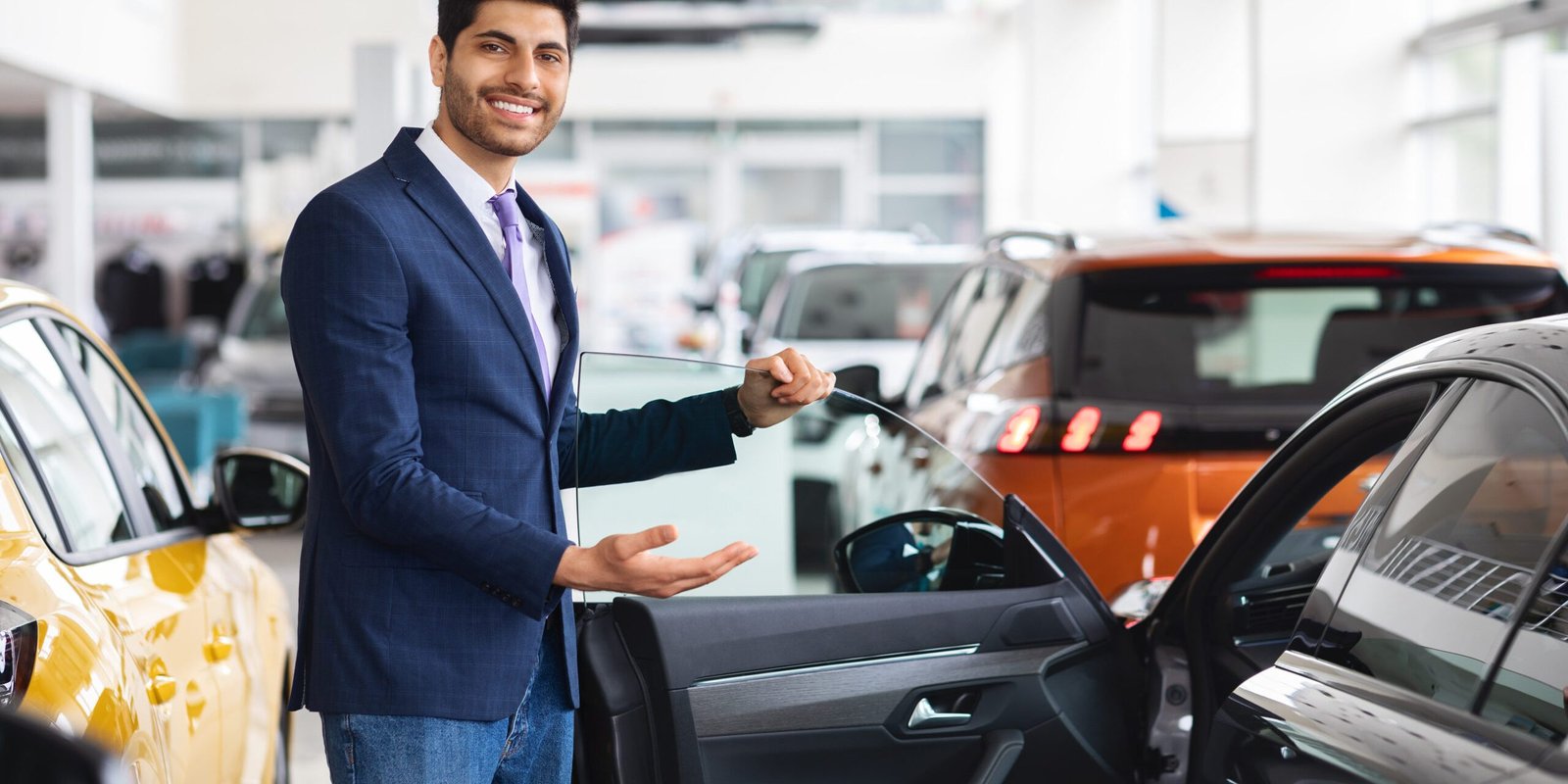 Cheerful young middle eastern man in formal outwear sales manager showing nice sports car, open black auto door and smiling, luxury automobile dealership salon interior, copy space